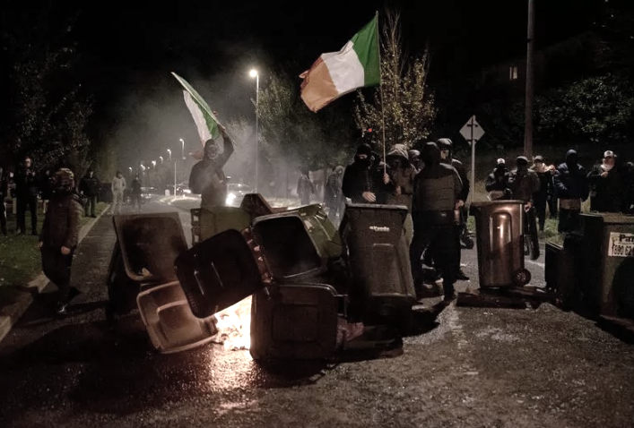 Night scene of masked protestors holding Irish tricolors and building a fire outside of Citywest Hotel.