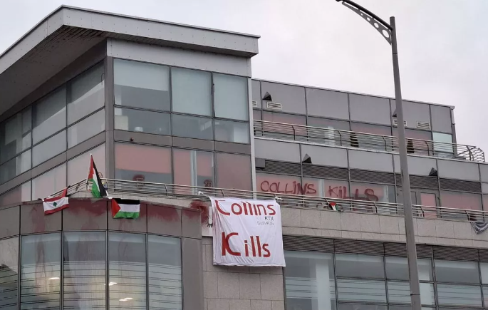 Cork Offices of Collins Aerospace with Palestine and Lebanon flags draped over railings next to a banner and grafitti saying Collins Kills.