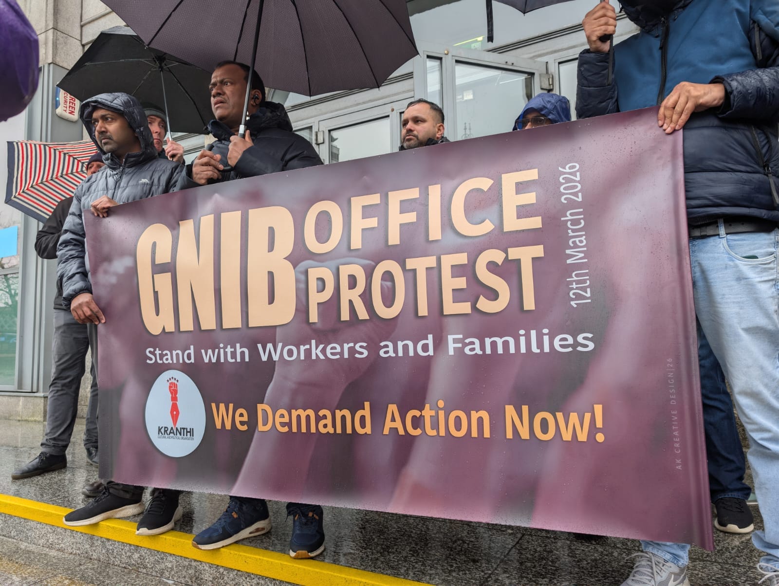 Workers standing in the rain at the GNIB Offices in Dublin with a banner that reads GNIB Office Protest 12th March 2026 - Stand with Workers and Families - We Demand Action Now! - Kranthi