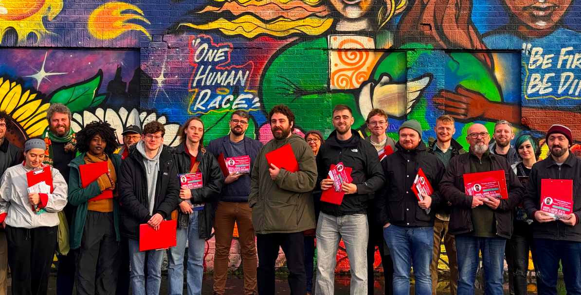 Eoghan Ó Ceannabháin, his campaign team, and supporters holding red cavassing clipboards standing in front of a brick wall covered with street art saying One Race Human Race. 