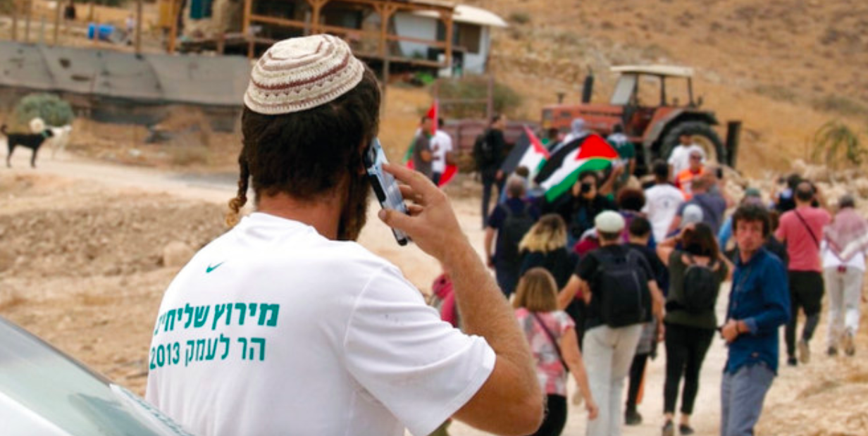 A man using a phone while watching marchers carrying Palestinian flags near a tractor.