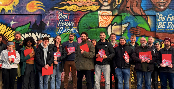 Eoghan Ó Ceannabháin, his campaign team, and supporters holding red cavassing clipboards standing in front of a brick wall covered with street art saying One Race Human Race. 