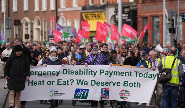 A 2026 Cost of Disability March in Dublin. Banner at head: Emergency Disability Payment Today. Heat or Eat? We Deserve Both. People Before Profit flags followed by other party flags