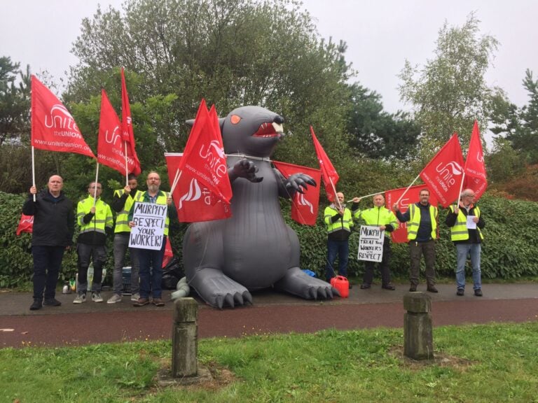 Unite the union members defend union rep at a protest with a giant inflatable rat and flags and hi-vis vests.